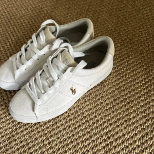 Vita sneakers från Polo Ralph Lauren - Snygga vita sneakers från Polo Ralph Lauren med klassisk design. Skorna har snörning, canvasmaterial och detaljer i mocka på hälen. Broderad logga på sidan och Polo-tryck på baksidan och sulan. Perfekta för en clean och stilren look.