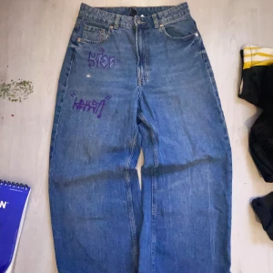 Blå wide jeans med lila graffiti - Säljer ett par blå Levi's jeans med bred passform och coola lila graffiti-prints på framsidan. Klassisk femficksmodell med knapp och dragkedja. Jeansen har hög midja och är tillverkade i robust denim. Perfekt för dig som vill sticka ut med din stil.
