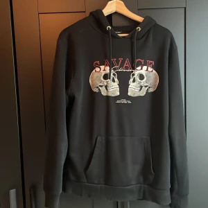 Svart hoodie med döskalletryck Savage - Svart hoodie från SikSilk med stort tryck av två döskallar och röd text 'SAVAGE' på bröstet. Klassisk känguruficka, snörning i huvan och mjukt material.