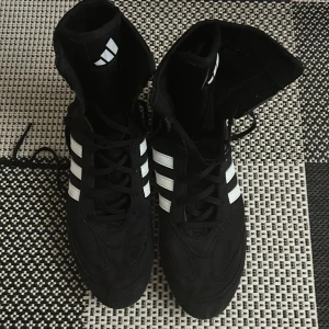 Svarta Adidas boxningsskor strl 44⅔ - Säljer ett par svarta Adidas boxningsskor med klassiska vita ränder på sidorna. Skorna har hög modell, snörning och platt vit sula med vågmönster för extra grepp. Materialet är syntet och mesh för lätthet och ventilation. Perfekt för dig som vill ha stil och funktion i ringen. 