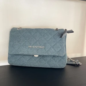 Jeansblå axelväska från Valentino - Snygg väska från Valentino i ljusblått jeanstyg med silverfärgad kedjerem och logga framtill. Väskan har en stilren, boxig form och lock med magnetlås. Oanvänt skick ✨Lägg gärna prisförslag