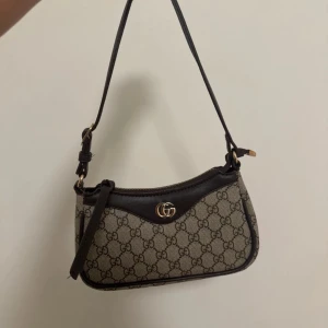  axelväska med GG-mönster - Snygg axelväska från  i beige och brunt med klassiskt GG-mönster över hela väskan. Väskan har detaljer i mörkbrunt skinn, justerbar axelrem och guldfärgad GG-logga framtill. Stängs med dragkedja och har en kompakt, stilren form.