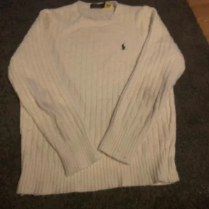 Vit stickad tröja från Polo Ralph Lauren - Vit stickad tröja från Polo Ralph Lauren med klassiskt broderat logotyp på bröstet. Tröjan har rund halsringning, ribbade muddar och långärmad passform. Perfekt för lager-på-lager under hösten och vintern.