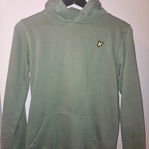 Grön hoodie från Lyle & Scott - Snygg grön hoodie från Lyle & Scott med klassisk gul logga på bröstet. Tröjan har huva, känguruficka och är tillverkad i mjuk bomull. Perfekt för chill dagar eller när du vill ha en enkel men stilren look.
