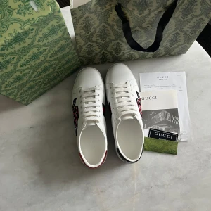 Gucci Ace sneakers med orm - Säljer ett par vita Gucci Ace sneakers i skinn med den ikoniska broderade ormen på sidan och röda samt gröna detaljer på hälen. Klassisk låg modell med snörning och rund tå. Kommer med originalkartong, dustbag och äkthetsbevis från vinted då dom är köpta for 4000. Byten kan vara intressant kom med frågor vid intresse😊