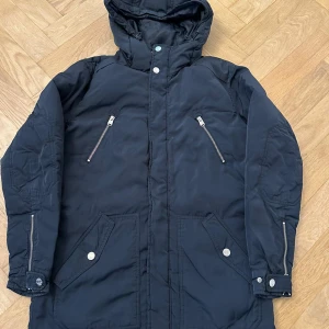 Mörkblå parkajacka med huva - Parkajacka i mörkblå färg med huva, dragkedjor på bröstfickor och ärmslut samt stora fickor framtill med knappar. Jackan har en rak passform och är perfekt för kyligare dagar.