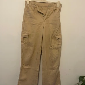 Beiga cargopants med raka ben - Snygga beiga cargopants med raka ben och klassiska cargofickor på sidorna. Byxorna har hällor för bälte och stängs med dragkedja och knapp. Perfekta för en avslappnad och trendig stil.