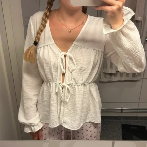 Vit blus med knyt från AWARE - Supersöt vit blus från AWARE med v-ringning och två knyt framtill. Långa ärmar med lätt volang vid ärmslut och en flowy, avslappnad passform. Tillverkad i ett mjukt, lätt material som känns luftigt och fräscht.