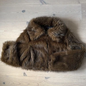 Brun pälsjacka från Pull&Bear XS - Snygg brun pälsjacka från Pull&Bear i storlek XS. Jackan har fluffig fuskpäls, bred krage och helsvart innerfoder. Perfekt för dig som vill sticka ut med en trendig och varm look under kyliga dagar. Modellen är midjekort. 