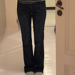Mörkblå bootcut jeans med fickor - Säljer ett par mörkblå jeans med bootcut passform och markerade sömmar längs benen. Jeansen har stora framfickor och dubbla knappar i midjan. Snygg och klassisk modell som är lätt att matcha till olika stilar.