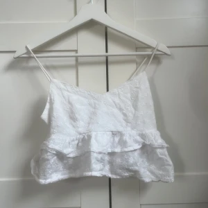 Vit volangtop med broderi - Söt vit linnetopp med tunna axelband och volang nertill. Toppen har broderade små blommor i tyget och en luftig, croppad passform. Perfekt för varma dagar och somriga outfits.