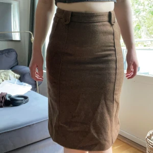 Brun ullkjol från Someday Soon - Snygg brun kjol från Someday Soon i klassisk rak modell med bälteshällor och diskreta sömmar framtill. Kjolen är gjord i mjuk ull och har en neutral brun ton som passar perfekt till stickade tröjor och boots. Perfekt för dig som gillar stilrena och tidlösa plagg.