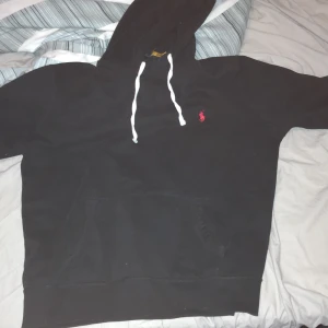 Äkta Svart hoodie från Ralph Lauren - Svart hoodie från Ralph Lauren med vit snörning och klassisk röd logga broderad på bröstet. Tröjan har huva, känguruficka och är tillverkad i mjuk bomull. Perfekt för en chill och stilren look.