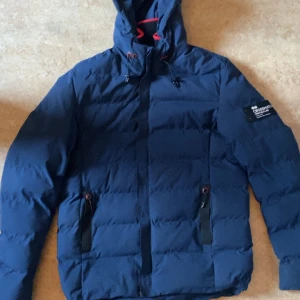 Blå pufferjacka från Crosshatch  - Säljer en marinblå pufferjacka från Crosshatch Black Label i storlek Large. Jackan har huva med snörning, svarta detaljer vid fickorna och patch på ärmen. Perfekt för kalla dagar med sin vadderade design och stilrena look.