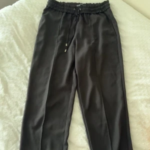 Svarta byxor med snörning från Zara - Svarta byxor från Zara med elastisk midja och snörning. Byxorna har en rak passform och är tillverkade i ett mjukt, lätt material som ger en avslappnad men ändå stilren look. Perfekta att matcha med en crop top eller hoodie.