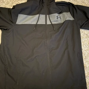 Svart vindjacka från Under Armour L - Svart vindjacka från Under Armour med grå panel över bröstet och logga. Jackan har huva med snörning, dragkedja framtill och två fickor. Meshfoder på insidan och loose fit för en avslappnad stil. Perfekt för blåsiga dagar.