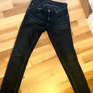 Svarta slim fit från Nudie Jeans - Snygga svarta slim fit från Nudie Jeans med klassisk femficksdesign och silverfärgade knappar. Jeansen har en tight passform och är tillverkade i stretchig denim för extra komfort. Perfekta för dig som gillar en stilren och modern look.