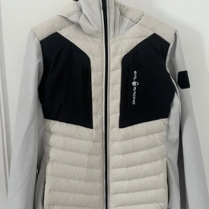 Sail Racing pufferjacka med huva - Snygg pufferjacka från Sail Racing i svart och ljusgrått med broderad logga på bröstet och patch på ärmen. Jackan har huva, två bröstfickor med dragkedja och quiltad design. Perfekt för kalla dagar och riktigt stilren look.