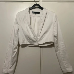 Vit croppad kavaj från Zoul - Snygg vit croppad kavaj från Zoul med klassisk krage och en knapp framtill. Jackan har lång ärm och är tillverkad i ett slätt tyg som ger en clean look. Perfekt för dig som vill ha en stilren och trendig touch till outfiten. Behöver bara strykas!