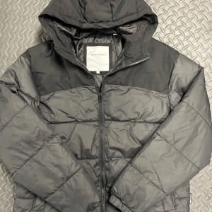Svart pufferjacka från Jack & Jones - Snygg svart pufferjacka från Jack & Jones med huva och dragkedja. Jackan har quiltad design och är tillverkad i syntetmaterial. Perfekt för kalla dagar när du vill hålla stilen och värmen. Oanvänd 
