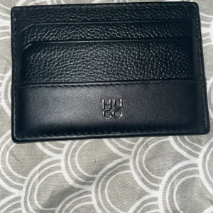 Svart kortfodral från Hugo - Stilrent svart kortfodral från Hugo i äkta skinn. Fodralet har flera kortfack och en snygg kombination av slät och präglad läderstruktur. Perfekt för dig som vill ha ett minimalistiskt och modernt accessoar med diskret logga framtill.