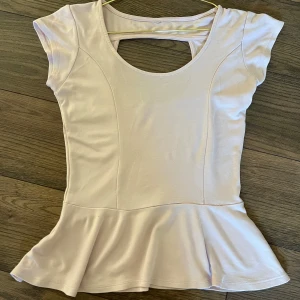 Ljusrosa peplum topp med cut out - Söt ljusrosa topp med peplumvolang och rund halsringning. Toppen har korta ärmar och en snygg cut out-detalj i ryggen. Tillverkad i mjukt och stretchigt material som ger en bekväm passform.