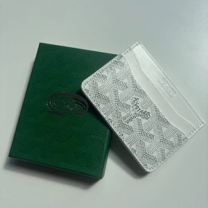 Vit kortfodral från Goy4rd - Supersnyggt vitt kortfodral från Goy4rd med deras ikoniska mönster i svart och grått. Slimmad design med flera kortfack och präglad logga framtill. Tillverkad i exklusivt skinn och levereras med original grön ask. Perfekt för dig som vill ha något stilrent och lyxigt i fickan. Tveka inte på att skicka om ni har några funderingar.