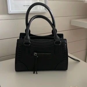 Svart handväska  - Svart handväska med både handtag och avtagbar axelrem. Väskan har snygga detaljer med flätad effekt på sidorna och en dragkedja framtill. Perfekt storlek.