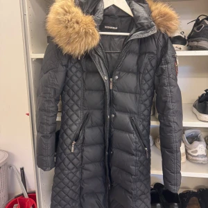 Svart lång dunjacka Rockandblue - Snygg svart lång dunjacka från Rockandblue med quiltad design och stor huva med fluffig päls. Jackan har dragkedjor på fickorna och längs framsidan, samt markerad midja för en schysst siluett. Perfekt för kalla dagar! Jag tycker att den är i bra skick!