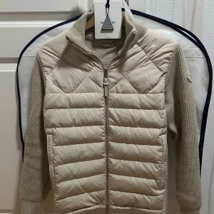 Moncler Cardigan  - Snygg beige dunväst från Moncler med quiltad framsida och ribbstickade ärmar och rygg. Hög krage, hel dragkedja framtill och Moncler-logga på ärmen. Perfekt lager-på-lager-plagg med exklusiv känsla och stilren design.