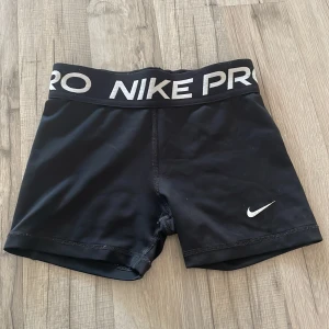 Nike Pro svarta träningstights shorts - Svarta Nike Pro shorts med bred resår i midjan där det står 'NIKE PRO' i vitt. Tight passform och tillverkade i Dri-FIT-material som andas. Perfekta för träning och sport. Klassisk Nike Swoosh-logga på benet.