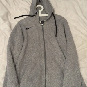 Grå Nike hoodie med dragkedja XL - Säljer en grå hoodie från Nike i storlek XL. Tröjan har hel dragkedja, huva med svarta snören och två fickor framtill. Perfekt för chill dagar eller när du vill ha en enkel och stilren look.