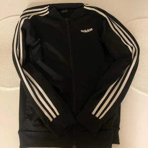 Svart Adidas ziptröja med vita ränder XS - Svart ziptröja från Adidas i storlek XS med klassiska vita ränder längs ärmarna och Adidas-logga på bröstet. Tröjan har hel dragkedja framtill och ribbade muddar. Perfekt för en sportig och avslappnad stil.