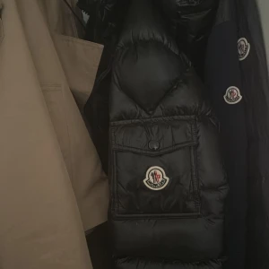 Svart Moncler dunjacka med ficka - Svart dunjacka från Moncler med klassisk logga på bröstet och stor ficka framtill. Jackan har glansig finish, dragkedja och quiltad design. Insidan har ett unikt serietidningsmönster och märkestaggar. Perfekt för dig som vill ha en riktigt snygg vinterjacka.