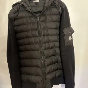 Svart Moncler cardigan - Svart cardigan från Moncler med quiltad front och huva. Jackan har stickade ärmar, dragkedja framtill och en ficka med Moncler-logga på ena ärmen. I princip helt ny, knappt använd, inga defekter. Pris kan diskuteras. Söker även byten, kom med förslag.