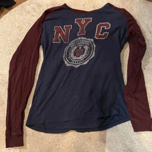 Blå långärmad NYC-tröja Dressmann - Snygg långärmad tröja från Dressmann i marinblått med vinröda ärmar. Stor NYC-print och varsity-tryck framtill ger en sportig collegekänsla. Skön passform och tillverkad i mjuk bomull, perfekt för dig som gillar avslappnad stil.