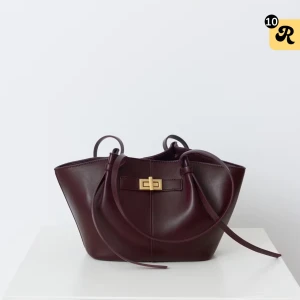 Mörkbrun axelväska i skinn med gulddetalj - Snygg mörkbrun axelväska i slätt skinn med stilren guldfärgad låsdetalj framtill. Väskan har två långa axelband och en rymlig, lätt utsvängd form som ger en modern vibe. Perfekt för dig som vill ha en elegant och praktisk väska med exklusiv känsla.