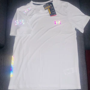 Vit Under Armour t-shirt med holografiska detaljer - Fräsch vit t-shirt från Under Armour med holografiska loggor och text på bröstet, ärmen och längs sidan. Tillverkad i lätt och ventilerande polyester, perfekt för träning. Klassisk passform med rund hals och korta ärmar.