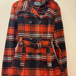 Rutig orange kappa från ONLY med huva - Snygg rutig kappa från ONLY i härlig höstfärg orange, svart och vitt. Jackan har avtagbar huva, bälte i midjan och dubbelknäppning med stora svarta knappar. Långärmad och i mjukt material som känns ulligt. Perfekt för dig som vill sticka ut i höst och vinter! Material: polyester.