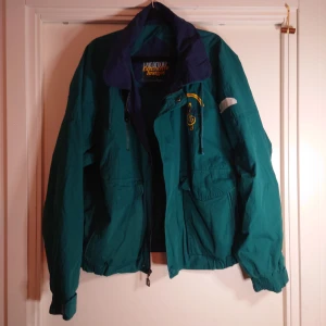 Grön vindjacka Dunbrooke L - Retro vindjacka från Dunbrooke i grön färg med mörkblå krage. Broderad detalj på bröstet med texten 'Wenger 50th Anniversary 1946-1996'. Jackan har dragkedja, stora fickor och elastiska muddar. Perfekt för dig som gillar vintage och unika plagg.