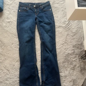 Blå bootcut jeans från Divided H&M - Snygga blå bootcut jeans från Divided H&M med klassisk femficksdesign och coola broderade detaljer på bakfickorna. Jeansen har orange kontrastsömmar och dragkedja framtill. Perfekta för dig som gillar en avslappnad men trendig look.