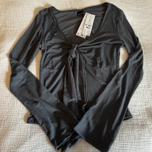 Grå långärmad topp med knyt från Gina Tricot - Supersnygg grå långärmad topp från Gina Tricot med djup v-ringning och knyt framtill. Toppens material är mjukt och stretchigt. (Slutsåld)                                   Oanvänd med lappen kvar.                                  Skickar ditt köp inom 24h!💘