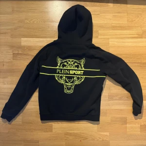 Svart hoodie från Plein Sport med tryck - Svart hoodie från Plein Sport med huva och snörning, hel dragkedja och två fickor med zip. På ryggen finns ett stort gult tigertryck och logga. Materialet är mjuk bomull och passformen är normal. Perfekt för dig som gillar sportig streetstyle.