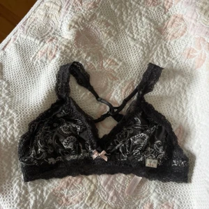 Svart spetsig bralette från Odd Molly - Supersnygg svart bralette från Odd Molly med spetsdetaljer och paisleymönster i vitt. Axelbanden är justerbara och korsas i ryggen. Söt liten rosett framtill och mjukt material som känns lyxigt mot huden. Perfekt för att ge outfiten en extra touch.