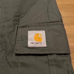 Gröna cargobyxor från Carhartt WIP - Snygga gröna cargobyxor från Carhartt Work In Progress med stora fickor på sidorna och justerbara spännen i midjan. Byxorna är tillverkade i slitstark bomull och har klassisk Carhartt-logga på benet. Perfekta för en streetwear-look.