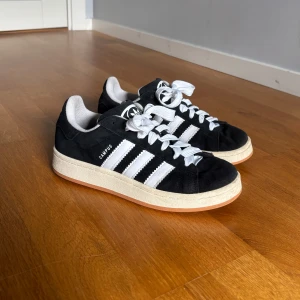 Adidas Campus svart/vit sneakers 38⅔ - Säljer dessa svarta Adidas Campus sneakers i mocka med klassiska vita ränder och vit sula. Skorna har rund tå, platt sula och vita snören🥰🥰 dom är använda men i väldigt bra sick och inga defekter💞