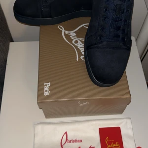 Christian Louboutin svarta sneakers mocka - Snygga svarta sneakers från Christian Louboutin i mocka med klassisk röd sula. Skorna har rund tå, svarta snören och broderad logga baktill. Kommer med originalkartong, dustbag och äkthetskort. Perfekta för dig som vill sticka ut med exklusiv stil.