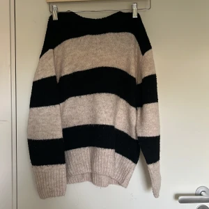 Randig stickad tröja från Gina Tricot - Mysig stickad tröja från Gina Tricot med breda svarta och beige ränder. Tröjan har en relaxed fit, ribbade muddar och lång ärm. Perfekt för kyliga dagar när du vill vara både bekväm och snygg.