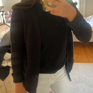 Svart stickad kofta med fickor - Mysig svart stickad kofta från laget 156 med grovstickat mönster och två praktiska fickor framtill. Koftan har lång ärm och bred ribbad kant längs öppningen och är perfekt att slänga på sig när det är kyligt. Jätte fin kofta som man kan ha över ett vanligt vit linne.