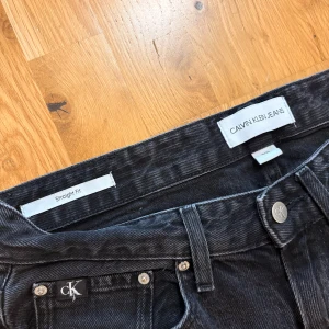 Svarta straight fit jeans Calvin Klein - Svarta jeans från Calvin Klein med straight fit och klassisk femficksdesign. Jeansen har diskreta CK-detaljer på fickan och knappen, och är tillverkade i ett robust jeanstyg. Perfekta för dig som gillar en clean och tidlös look. 